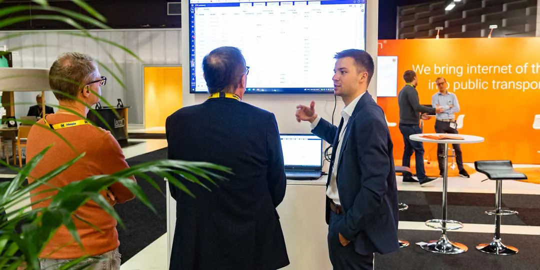 Bli-utställare_2L5A0021_1080x540 Tre personer står vid en mässmonter och samtalar framför en stor skärm som visar data i ett digitalt gränssnitt. En person i mörk kostym gestikulerar mot skärmen medan de två andra lyssnar. I bakgrunden syns en orange montervägg med text om internetlösningar för kollektivtrafik, samt flera ståbord och besökare. Gröna växter ramar in förgrunden och miljön präglas av en modern mäss- eller utställningshall.