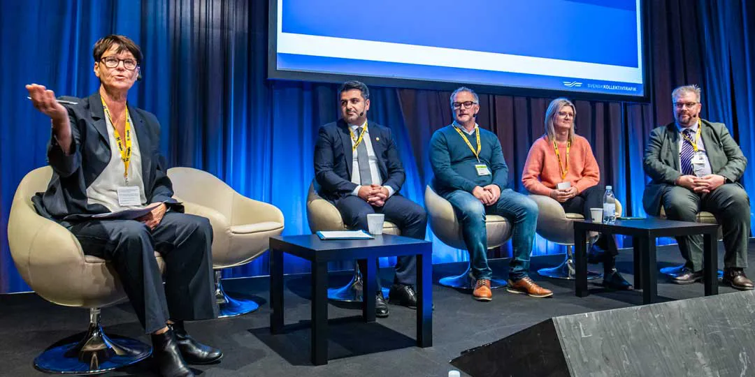 Moderator_2L5A0750_1080x540 Paneldiskussion på en konferensscen med fem deltagare sittande i fåtöljer framför en blå bakgrund och en stor presentationsskärm. En person längst till vänster håller i papper och gestikulerar mot publiken, medan övriga paneldeltagare sitter på rad bredvid. Framför deltagarna står små bord med vattenflaskor och anteckningsmaterial. Samtliga bär namnbricka på gul rem och scenen är upplyst med blått ljus.