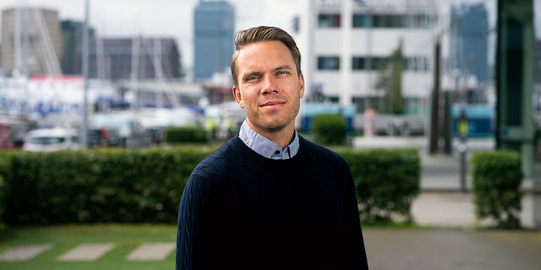 En person i mörk tröja och ljus skjorta står utomhus framför en modern stadsmiljö med kontorsbyggnader och en marina i bakgrunden. Personen tittar in i kameran.