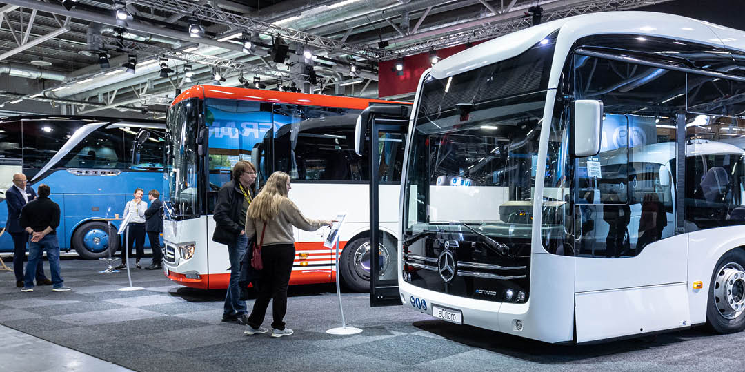 PT_puffbild_tavling_Vagvisaren_1080x540px Flera moderna bussar visas upp på mässan Persontrafik där besökare tittar närmare på fordonen och samtalar i utställningshallen.