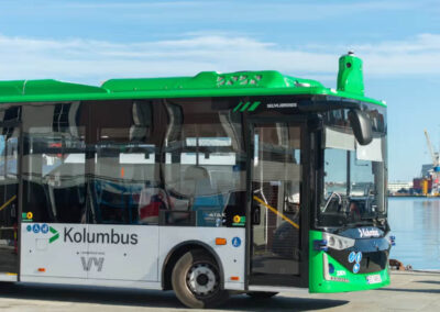 En grön och vit självkörande buss från Kolumbus står parkerad vid kajen i en hamn. Bussen är fotograferad snett framifrån med vatten, fartyg och hamnkranar i bakgrunden under en klarblå himmel. På bussens front visas texten "Selvkjørende", som markerar att den kör autonomt.