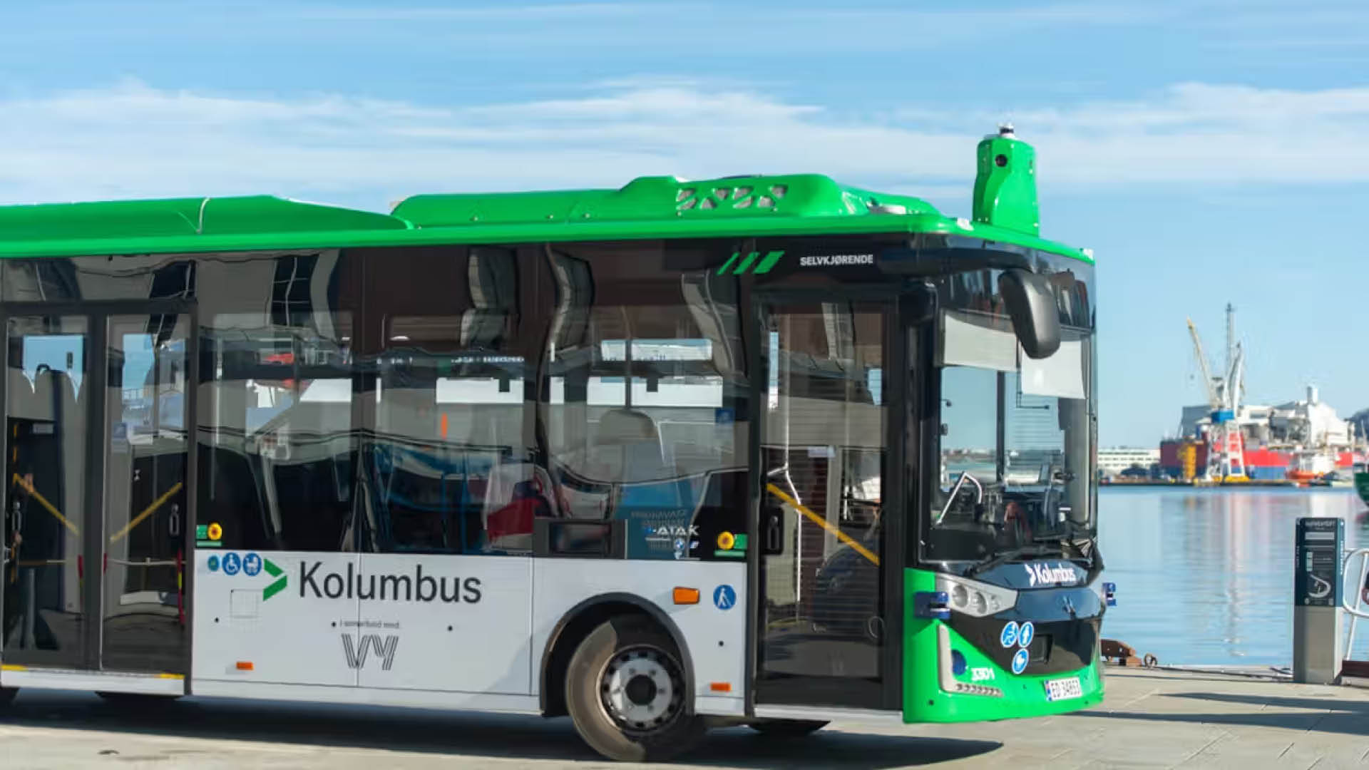 PT_toppbild_autonoma_fordon_NY_1920x1080px En grön och vit självkörande buss från Kolumbus står parkerad vid kajen i en hamn. Bussen är fotograferad snett framifrån med vatten, fartyg och hamnkranar i bakgrunden under en klarblå himmel. På bussens front visas texten "Selvkjørende", som markerar att den kör autonomt.