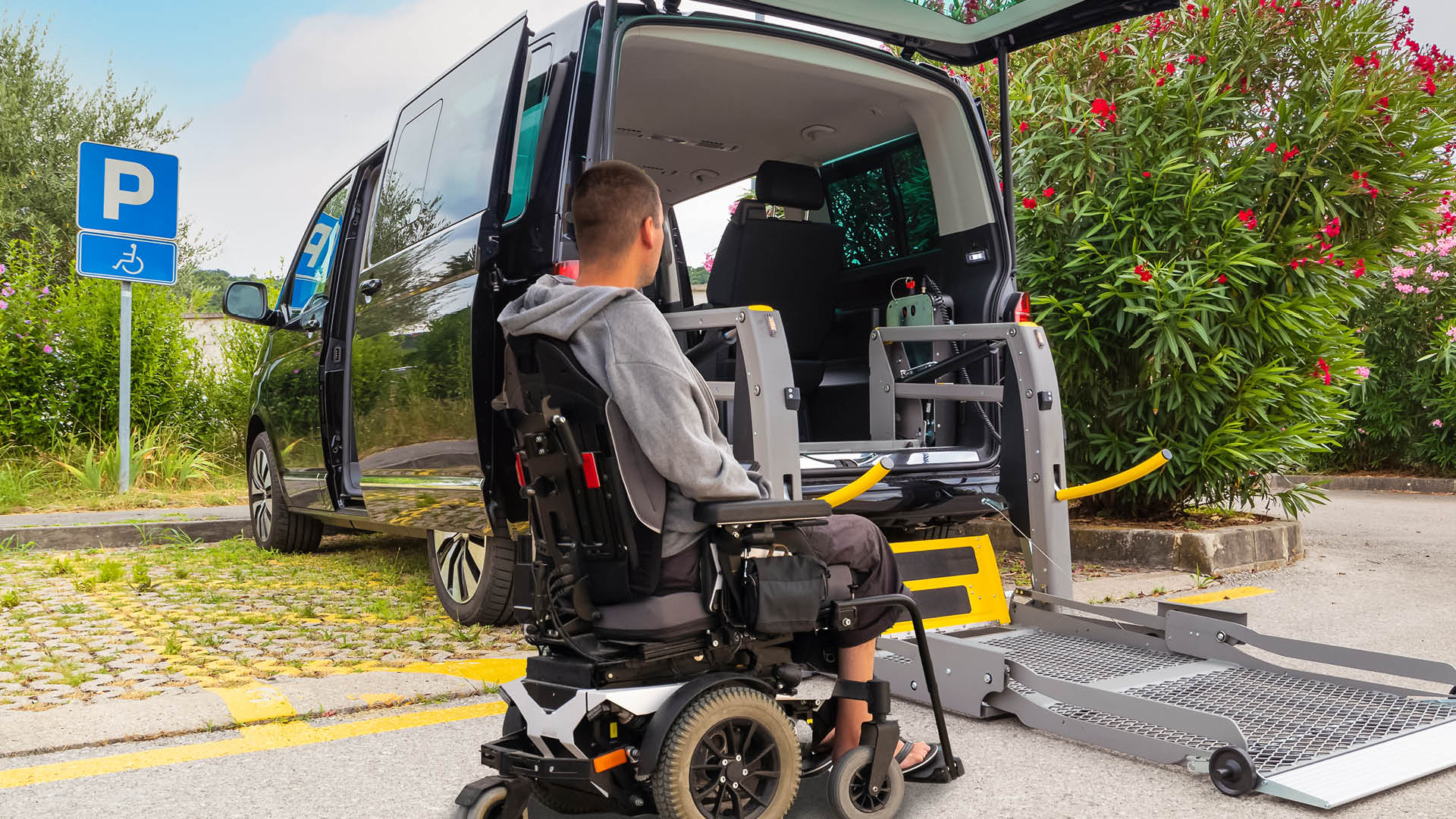 Fråga experterna på Persontrafik_1920x1080px Person i elrullstol framför en tillgänglighetsanpassad bil med utfälld ramp vid en parkeringsplats för personer med funktionsnedsättning.