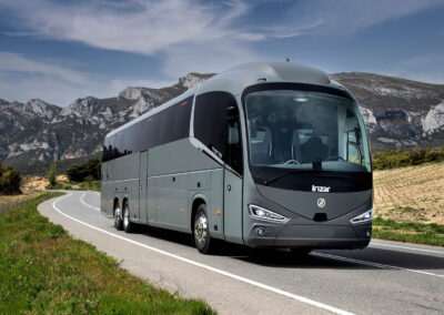 Grå Irizar turistbuss kör på en landsväg med bergslandskap i bakgrunden.