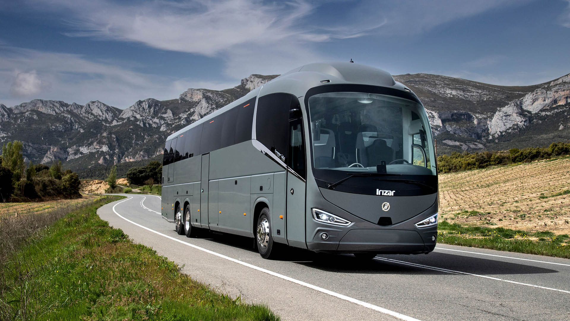 Grå Irizar turistbuss kör på en landsväg med bergslandskap i bakgrunden.