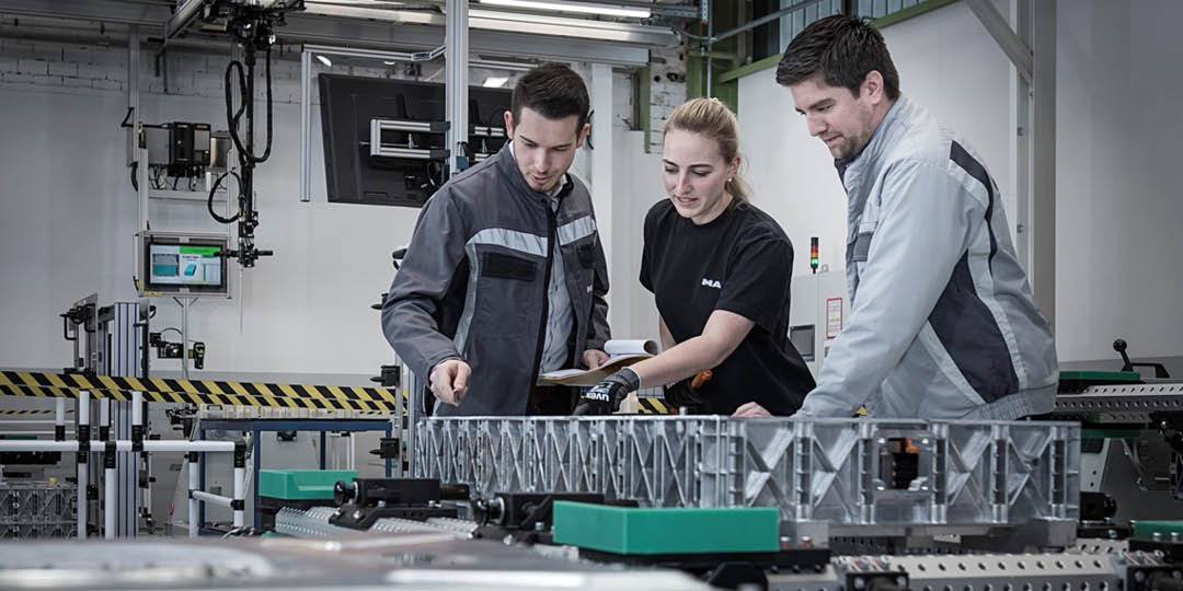 MAN_manniskor_1080x540px Three MAN employees inspecting a component together in a modern production facility.