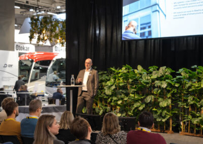 En föreläsare presenterar på scen inför en sittande publik på Persontrafik, med bussar och utställarmontrar i bakgrunden.