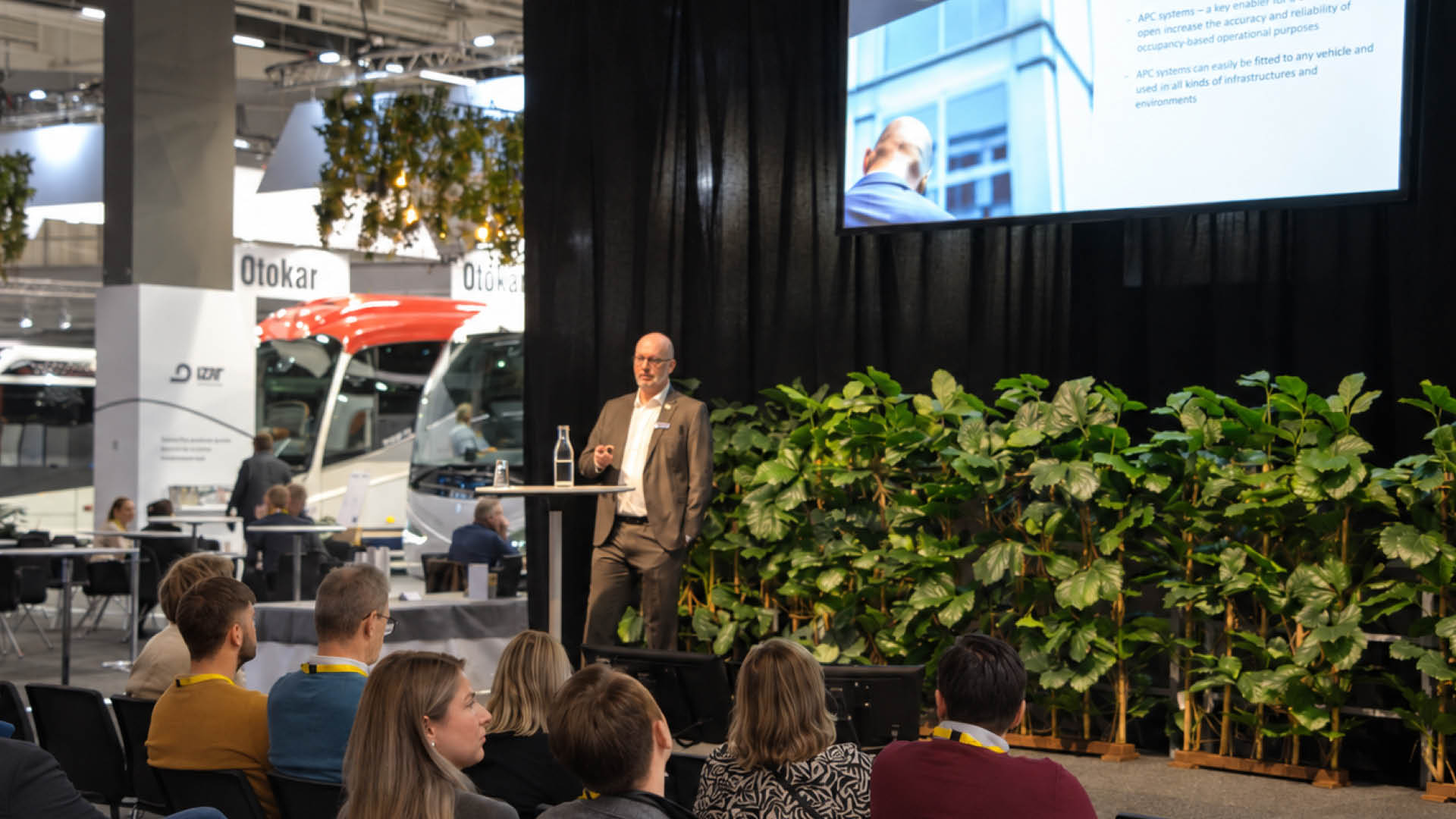 En föreläsare presenterar på scen inför en sittande publik på Persontrafik, med bussar och utställarmontrar i bakgrunden.