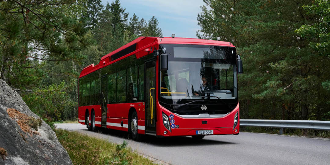 Scania_1080x540px Red Scania city bus travelling along a road through a wooded area.