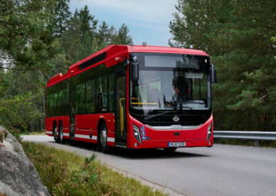 Röd Scania stadsbuss kör på en väg genom ett skogsområde.