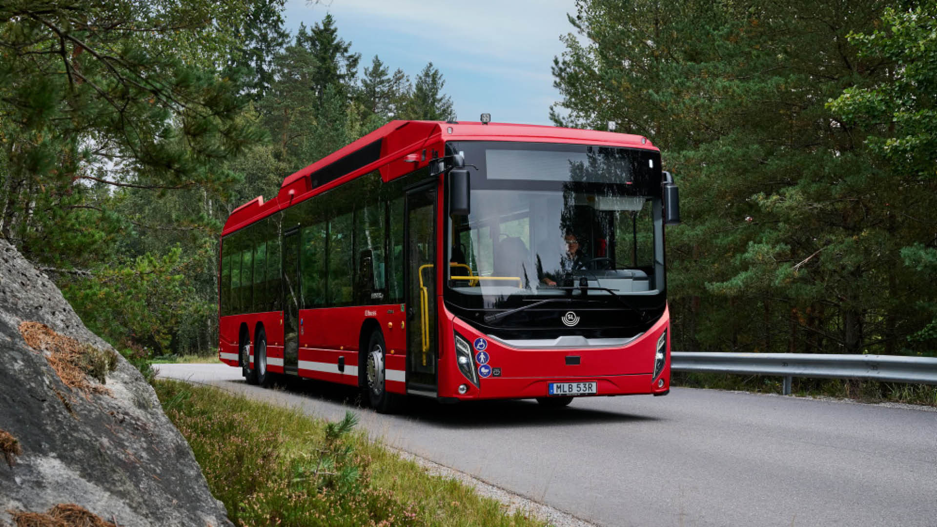 Röd Scania stadsbuss kör på en väg genom ett skogsområde.